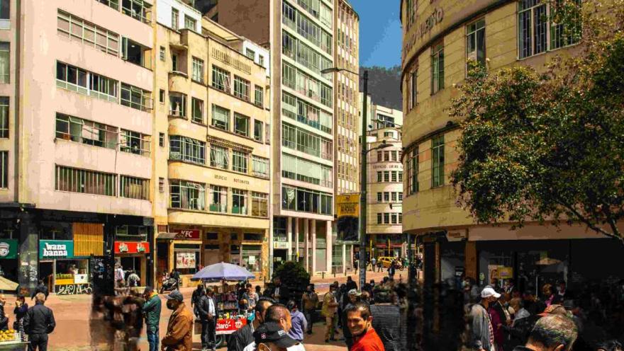 A busy city street with people walking and standing, surrounded by buildings and traffic.