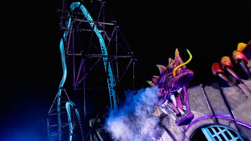  Atracción de montaña rusa iluminada de noche en un parque de diversiones. En primer plano se ve la escultura de un dragón morado que expulsa humo, mientras detrás se eleva la estructura metálica de la montaña rusa con rieles iluminados contra el cielo oscuro.
