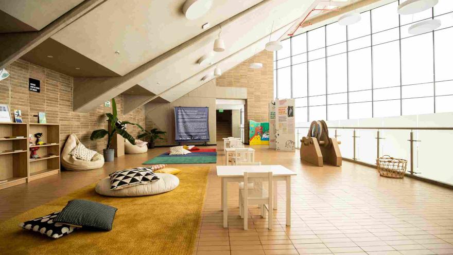  Espacio interior luminoso acondicionado para actividades infantiles, con mesas y sillas pequeñas, cojines sobre alfombras y estanterías con libros y materiales. El lugar cuenta con grandes ventanales que dejan entrar luz natural y paredes de ladrillo que crean un ambiente cálido y educativo.
