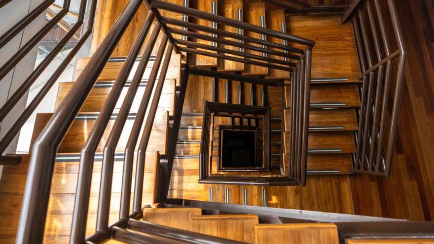Vista hacia abajo de una escalera interior de madera con barandas metálicas oscuras que forman una figura geométrica en espiral hacia el centro. Los peldaños de madera iluminados por luz natural crean un patrón cálido y simétrico que desciende varios niveles del edificio.