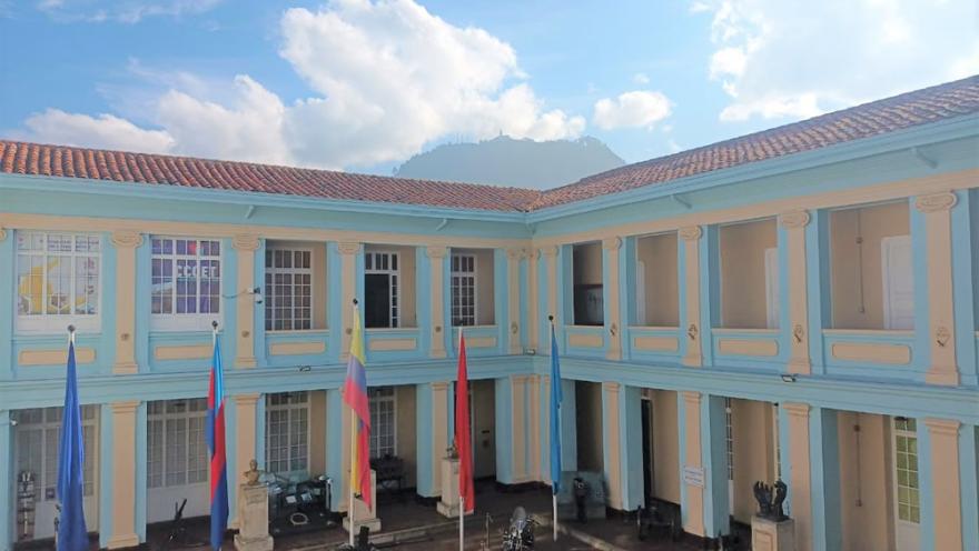  Patio interior de un edificio histórico de dos pisos con columnas y balcones, banderas izadas en el centro y piezas militares exhibidas, con montañas visibles al fondo.