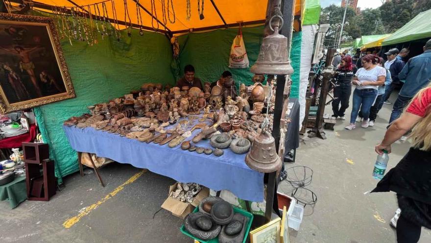An open-air market stall under a yellow tent displays antique and handcrafted objects such as figurines, utensils, stones, and decorative pieces on a covered table. Vendors are serving customers in the background, and visitors are browsing to one side.
