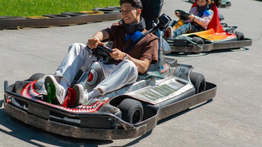 Dos personas conducen karts en una pista al aire libre en un parque de diversiones. En primer plano, un joven sonríe mientras maneja su kart gris, seguido por otra persona en un kart naranja. Alrededor de la pista se observan llantas de protección y césped verde.