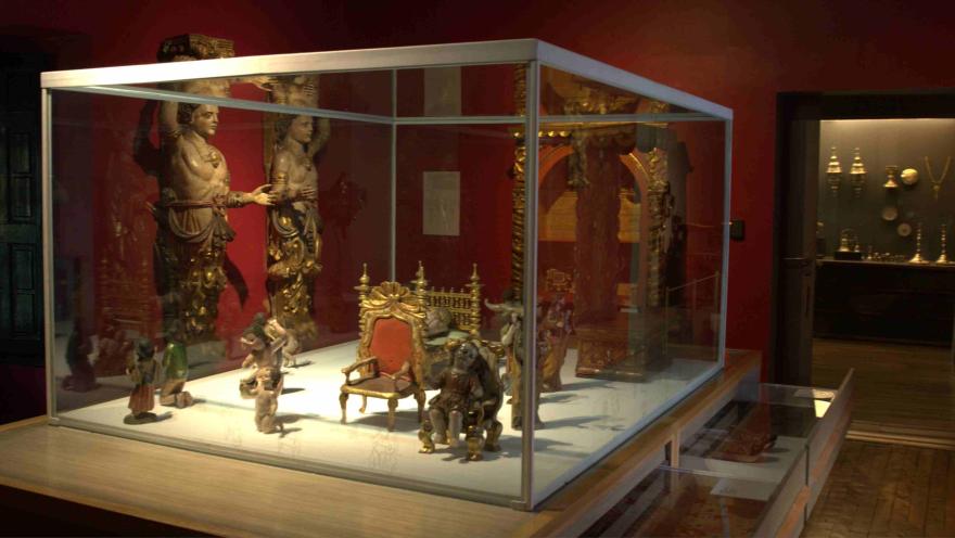Vitrina de museo con esculturas y objetos decorativos antiguos, incluyendo pequeñas figuras, un trono en miniatura y piezas talladas, en una sala de paredes rojas con iluminación tenue.