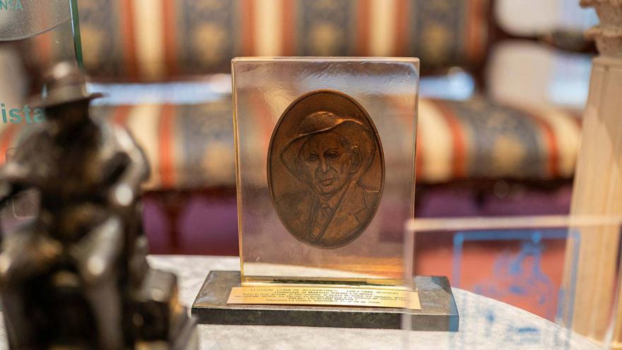  Objeto conmemorativo exhibido en una vitrina, compuesto por una placa con relieve de un rostro masculino sobre una base con inscripción. La pieza tiene un acabado en tonos dorados y está rodeada de otros elementos decorativos y reconocimientos, en un ambiente interior tipo museo o sala de exposición.
