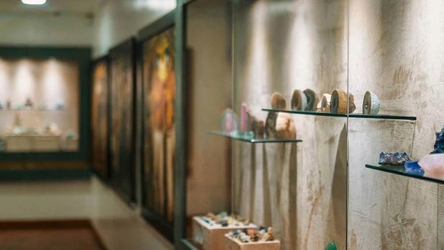  Interior de una sala de museo con vitrinas de vidrio donde se exhiben minerales y piedras preciosas sobre repisas transparentes. A lo largo del pasillo se observan más vitrinas y paneles expositivos iluminados que muestran diferentes piezas de la colección.