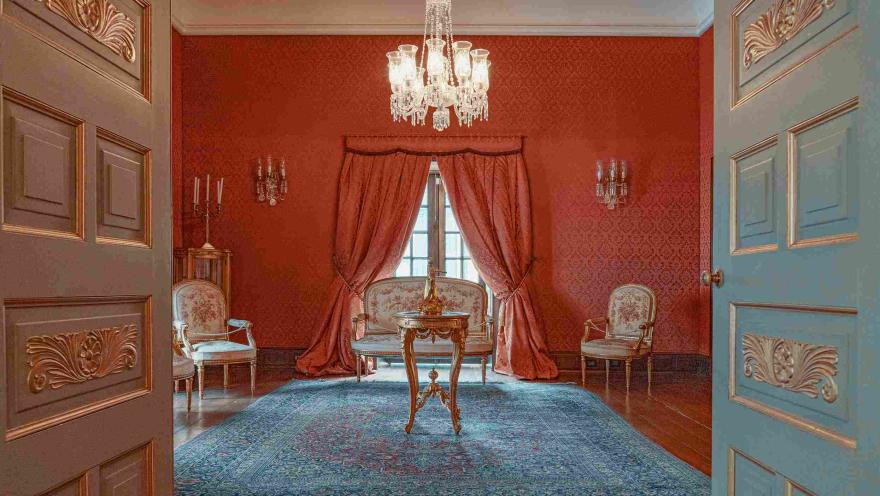  Vista de un elegante salón de estilo clásico en tonos rojos, con paredes decoradas y cortinas pesadas enmarcando una ventana central. En el centro hay una mesa de madera tallada sobre una alfombra azul, rodeada de sillas tapizadas de estilo antiguo. Un candelabro de cristal cuelga del techo, y la escena se observa desde una puerta abierta que enmarca el espacio con detalles ornamentales.