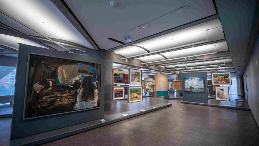Vista interior de la Sala de Arte Bancolombia, un espacio expositivo moderno donde se exhiben diversas pinturas de gran formato y obras enmarcadas sobre paneles y muros. La sala amplia, con iluminación dirigida desde el techo, permite recorrer la exposición y apreciar las obras desde distintos ángulos en un ambiente contemporáneo.