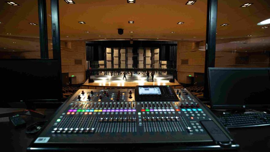  Vista desde la cabina técnica de un teatro, donde se observa una consola de sonido con botones y luces de colores en primer plano. Al fondo se ve el escenario iluminado y parte del auditorio con filas de asientos, mientras algunas personas caminan sobre el escenario durante un ensayo o preparación técnica.