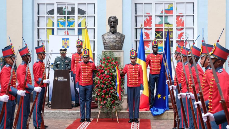 Ceremonia militar con guardia de honor uniformada frente a un busto conmemorativo, acompañada por banderas y una ofrenda floral en el patio de un edificio histórico.