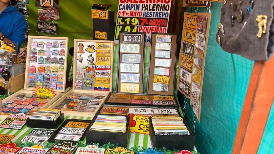 A market stall displaying decorative plaques and colorful souvenirs inspired by popular names, places, and expressions. The items are arranged on tables and vertical panels, with eye-catching signs advertising special offers.