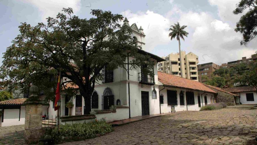Casa colonial blanca con techos de teja roja y un pequeño campanario, rodeada de árboles y un patio empedrado, con edificios modernos visibles al fondo.
