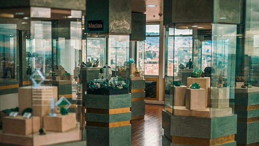 Sala de exposición de un museo con varias vitrinas de vidrio que exhiben esmeraldas y minerales sobre pedestales. Las piezas están organizadas en columnas dentro de un espacio iluminado, y al fondo se observa una ventana con vista a la ciudad.