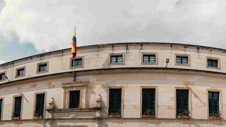 Vista panorámica de la fachada de un edificio histórico de estilo colonial con forma curva, construido en piedra clara. En el centro ondea la bandera de Colombia sobre la entrada principal, y a lo largo de la fachada se alinean ventanas con marcos oscuros y jardineras con flores. El cielo está parcialmente nublado, aportando un ambiente sobrio y patrimonial.