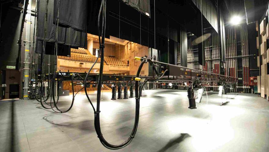  Vista desde el escenario de un teatro hacia el auditorio, donde se observan las graderías con asientos y la infraestructura técnica del escenario. En primer plano aparecen cables, estructuras de iluminación y sistemas de montaje escénico colgando del techo.