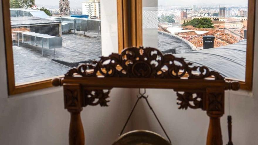  Interior de una habitación con una mesa decorativa de madera tallada y un pequeño gong colgante. Al fondo, a través de dos ventanas, se observa una vista del centro de la ciudad con techos de teja, edificios y una torre de iglesia sobresaliendo en el paisaje urbano.