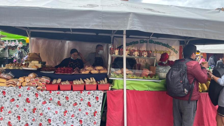 A food stall in an open-air market, with a white tent and covered tables displaying breads, cold cuts, and ready-to-eat dishes. Several people are serving customers at the front. The atmosphere is busy and dynamic, typical of an urban food fair.