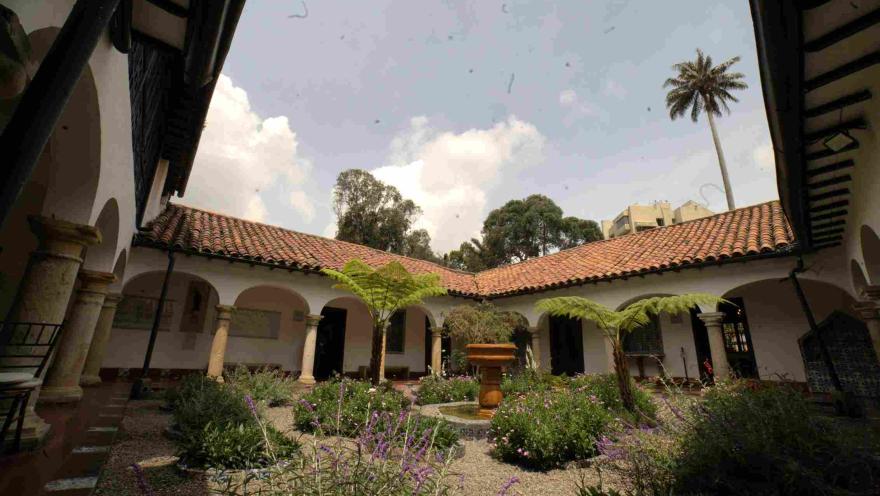  Patio interior de una casa colonial con corredores de arcos y columnas, techos de teja roja y un jardín central con plantas, flores y una fuente de piedra rodeada de vegetación.