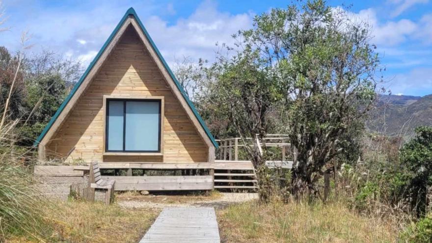 Casa en madera como una cabaña en forma de triángulo en medio de un bosque frondoso. En la casa hay una ventana cerrada y un camino de madera.
