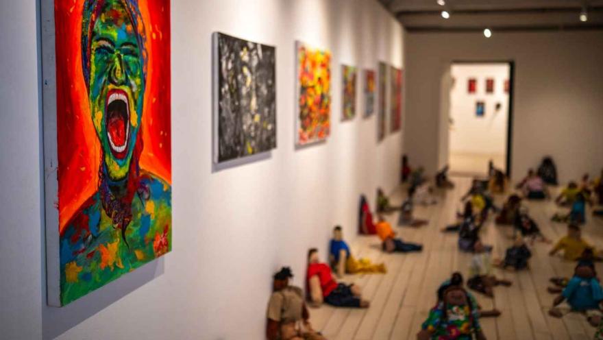 Interior de una galería de arte con varias pinturas coloridas colgadas en una pared blanca. En primer plano destaca un cuadro de una figura humana gritando en tonos intensos de rojo, verde y azul. A lo largo de la sala, en el piso, hay varias figuras humanas sentadas o recostadas como parte de una instalación artística, mientras al fondo se observa otra sala de exposición iluminada.
