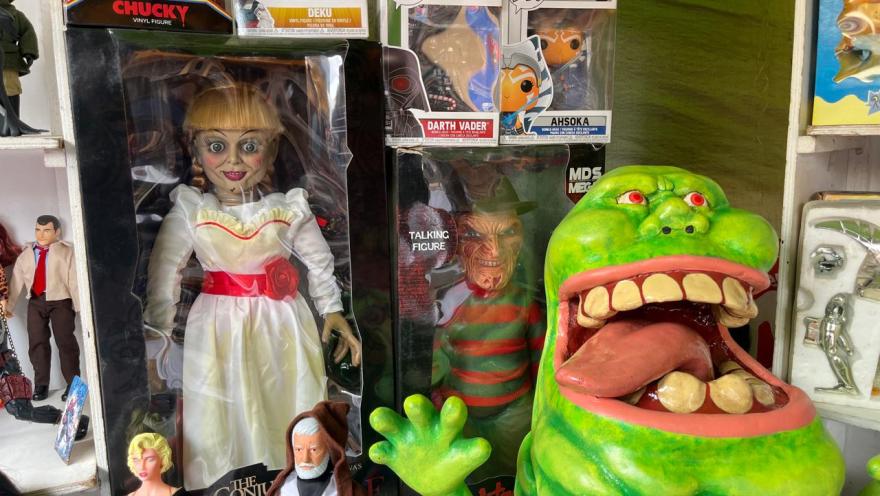 A shop window or stall displaying collectible pop culture figures, including action figures from movies and TV series, such as horror and science fiction characters. Boxes of Funko Pop-type figures and other detailed pieces are visible, along with a large, cartoonish green figure with its mouth open. The display showcases a varied and eye-catching collection for fans.