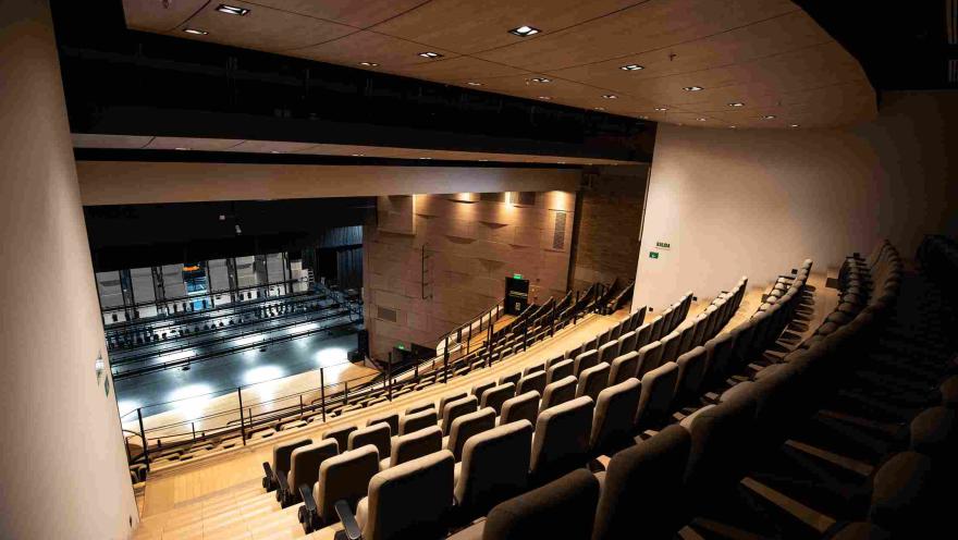 Vista lateral del interior de un teatro moderno con filas de asientos acolchados en gradería que descienden hacia el escenario. El espacio cuenta con iluminación cálida en el techo y barandas negras, mientras al fondo se observa el escenario con su estructura técnica.