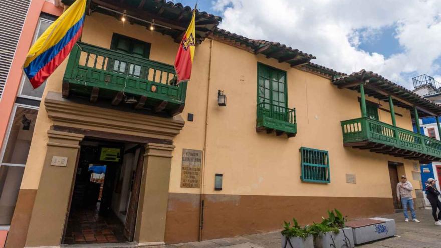 Fachada de una casa colonial de dos pisos con paredes color crema, balcones y ventanas de madera pintadas de verde. Sobre la entrada cuelgan las banderas de Colombia y de Bogotá. El edificio tiene techo de tejas de barro y una placa conmemorativa en la pared, mientras algunas personas caminan por la acera frente al lugar.