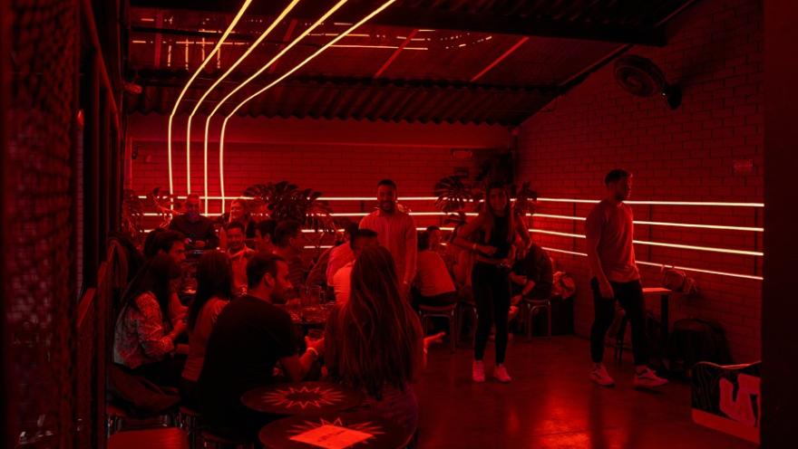  Interior de Tejo La Embajada concurrido, iluminado por una luz de neón roja vibrante, que muestra a personas socializando en mesas.