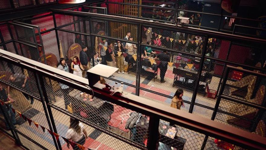 Vista elevada de un amplio espacio de eventos industrial con estructuras de malla metálica y una gran multitud de asistentes.