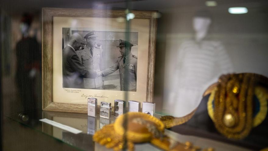  Vitrina de museo con objetos históricos de carácter militar o naval. En el centro se observa una fotografía en blanco y negro enmarcada, donde varios oficiales estrechan la mano, acompañada de pequeños objetos metálicos exhibidos en fila. En primer plano, desenfocado, se aprecia un elemento dorado decorativo, mientras la iluminación tenue resalta el valor histórico de la escena.