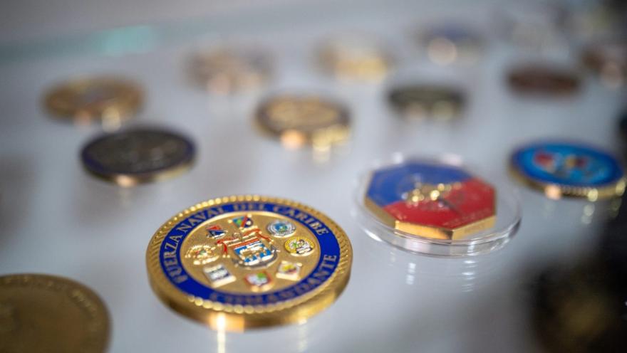Primer plano de una vitrina con medallas y monedas conmemorativas de estilo militar o naval. En el centro destaca una pieza dorada con detalles en azul y escudos, mientras otras insignias se distribuyen alrededor, algunas dentro de estuches transparentes.