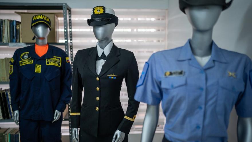 Tres maniquíes exhiben uniformes de la Armada en un espacio interior tipo museo. En el centro destaca un uniforme formal oscuro con botones dorados y gorra, mientras a los lados se observan otros trajes: uno azul con insignias y otro de tono claro con gorra. Al fondo hay estanterías con libros y una ventana con persianas que ilumina suavemente la escena.