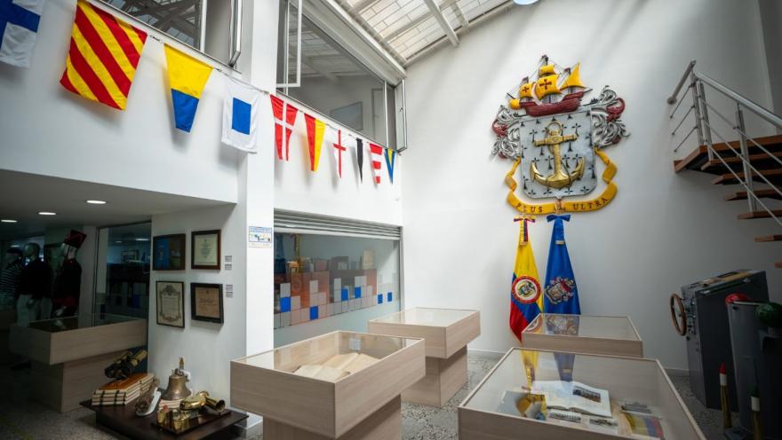  Interior de un museo con temática naval, iluminado por luz natural desde el techo. Se observan vitrinas de madera con documentos y objetos históricos, banderines marítimos colgados en la parte superior y, en una pared, un gran escudo con ancla y la frase “Plus Ultra”. A un lado están las banderas de Colombia y otra institucional, junto a una escalera metálica que conduce a un nivel superior.