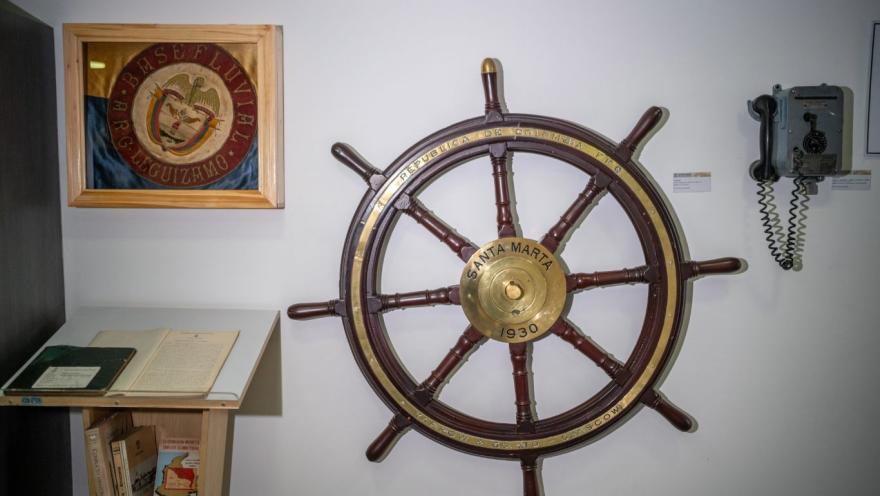 Exhibición en un museo con un gran timón de barco de madera y metal montado en la pared, con la inscripción “Santa Marta 1930”. A su lado hay un teléfono antiguo y, a la izquierda, un pequeño mueble con libros y documentos. Sobre este, se observa un cuadro con un escudo histórico, todo en un espacio iluminado que resalta el carácter naval y patrimonial de la muestra.