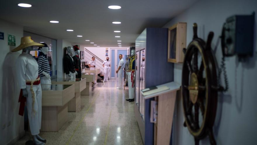  Pasillo interior de un museo o espacio expositivo con temática naval. A ambos lados se observan vitrinas y maniquíes vestidos con uniformes marítimos tradicionales, mientras al fondo varias personas con uniforme blanco conversan cerca de una escalera. En primer plano, a la derecha, destaca un timón de barco colgado en la pared, y la iluminación es moderna y uniforme, creando un ambiente ordenado y educativo.