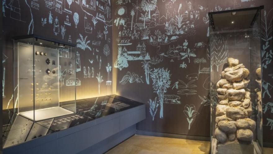  Interior de una sala de museo con iluminación tenue. En vitrinas de vidrio se exhiben objetos antiguos, como piezas metálicas y una estructura de piedras apiladas. Las paredes oscuras están decoradas con ilustraciones dibujadas en líneas blancas que representan paisajes, plantas, animales y escenas culturales, creando un ambiente histórico y educativo.
