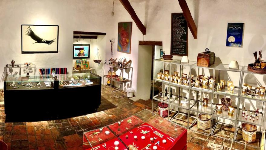 The interior of a craft shop with a brick floor and white walls, decorated with wooden beams on the ceiling. In the center and on the left are glass display cases showcasing jewelry and small handmade items. In the background, metal shelves display jars, natural products, and woven baskets.
