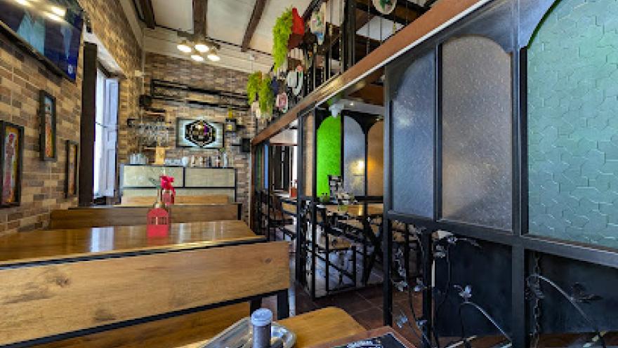 Interior de un restaurante de estilo rústico y acogedor, con paredes de ladrillo a la vista y vigas de madera en el techo. El espacio cuenta con mesas y bancas de madera, una barra al fondo y una estructura de dos niveles con barandas metálicas.