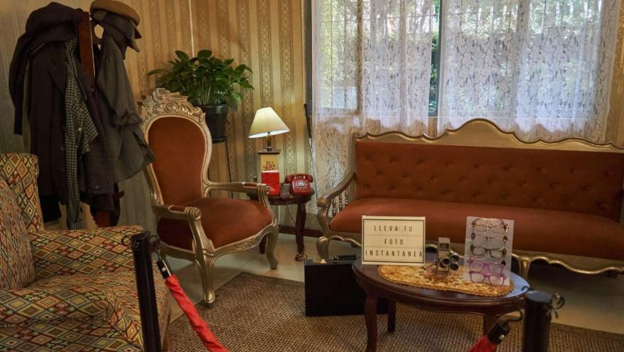 La imagen muestra un acogedor espacio interior de estilo vintage, con muebles clásicos como un sofá y sillones tapizados en tonos cálidos. Sobre una mesa se observan objetos antiguos, como un teléfono rojo y una cámara, junto a un letrero que invita a tomarse una foto.