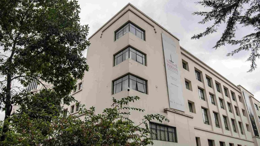 La imagen muestra un edificio de estilo sobrio y elegante, con fachada en tonos claros y grandes ventanas en esquina que aportan modernidad. Está rodeado de árboles y vegetación, lo que le da un ambiente tranquilo.