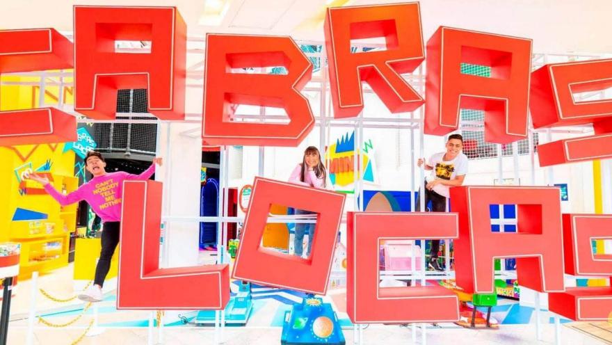 Three people pose and play among large red letters spelling "CABRAS LOCAS" in a colorful play area.