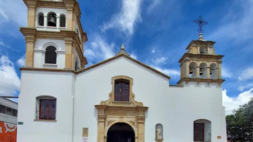 La imagen muestra la fachada de una iglesia de estilo colonial, de paredes blancas y detalles en tonos beige y ocre que resaltan su arquitectura. El edificio se compone de un cuerpo central con una entrada principal enmarcada por un arco ornamentado, sobre el cual se observa una ventana con reja y molduras decorativas. 
