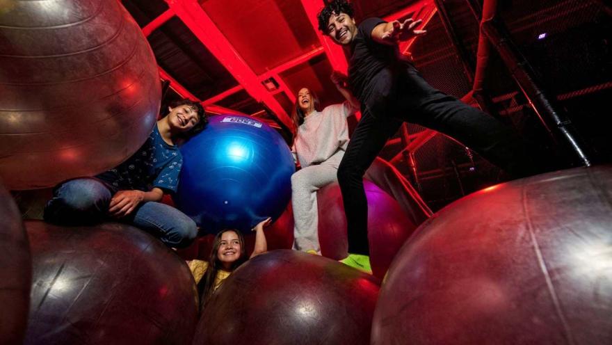 Cuatro personas sonríen y juegan sobre grandes pelotas de ejercicio en un espacio interior iluminado con luces rojas.