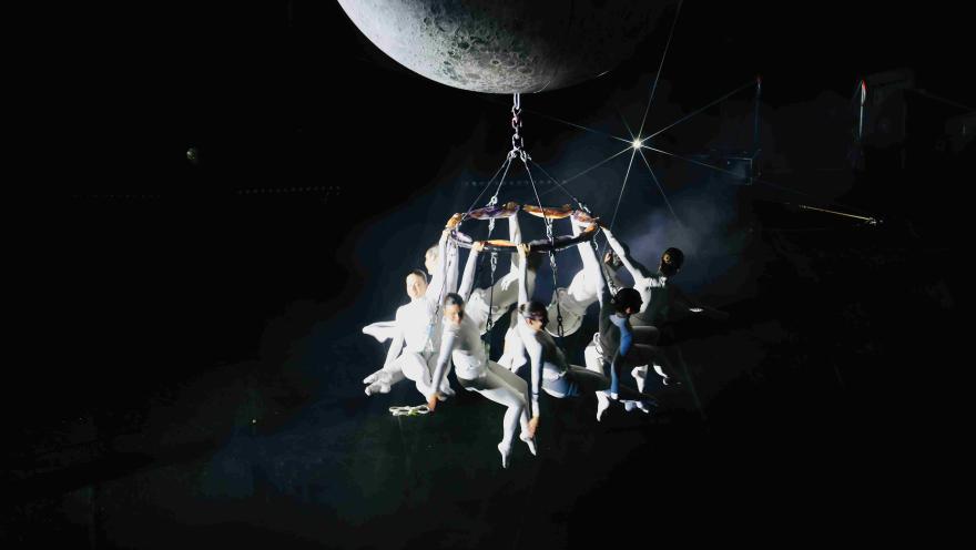 Escena artística en un espacio oscuro: varios acróbatas vestidos de blanco están suspendidos en una estructura colgante bajo una gran luna, iluminados por un foco que resalta la atmósfera teatral y aérea.
