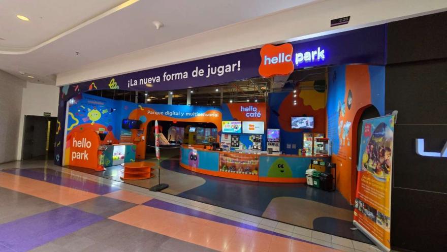 Entrada de “Hello Park” en un centro comercial, con fachada colorida, mostrador de atención, pantallas digitales y letreros que anuncian un parque multisensorial y cafetería.