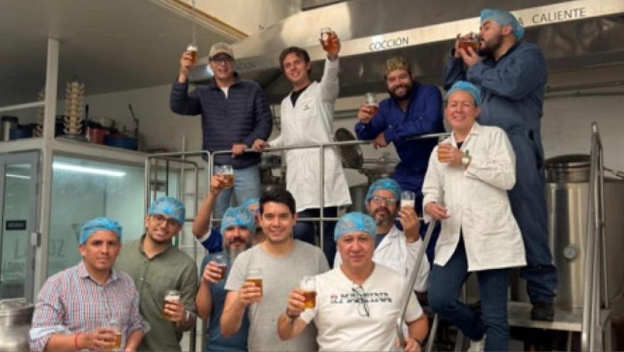 Grupo de once personas en una planta de producción, posando y brindando con vasos de cerveza. La mayoría lleva cofias o gorros de higiene y algunos visten batas blancas, lo que sugiere un entorno de elaboración de alimentos o bebidas.