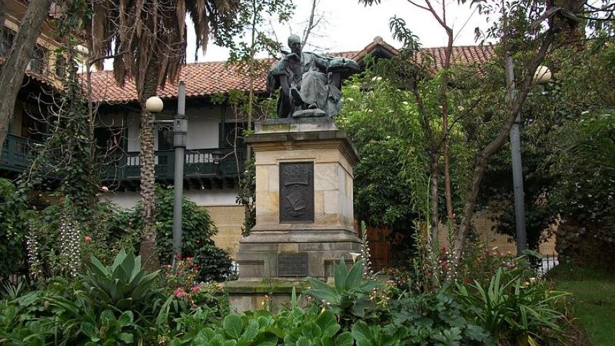 Estatua sobre pedestal en un jardín rodeado de árboles y plantas frente a una casa de estilo tradicional.