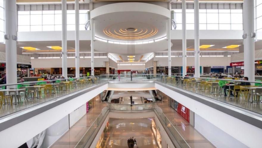 La imagen muestra el interior de un centro comercial amplio y luminoso, con una arquitectura moderna dominada por columnas blancas y grandes ventanales que dejan entrar abundante luz natural. En el nivel superior se observa una zona de comidas con mesas y sillas ocupadas por visitantes, mientras que en el centro se abre un vacío que conecta visualmente con los pisos inferiores mediante escaleras eléctricas.