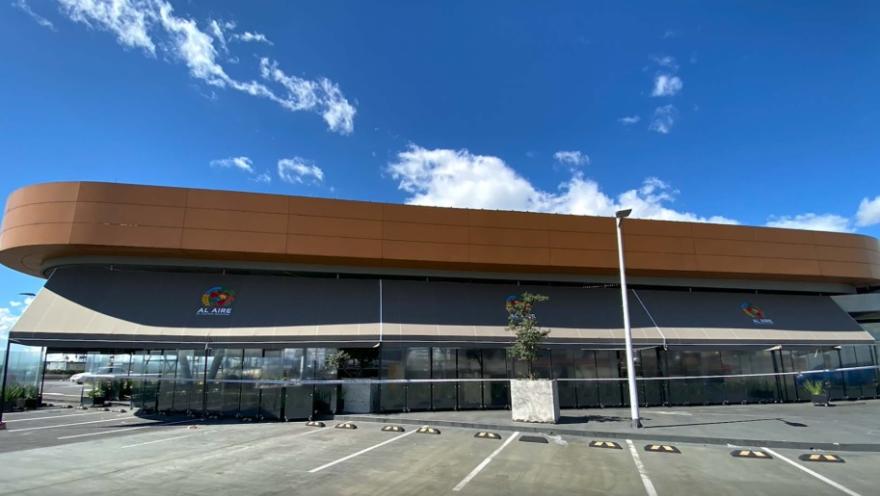La imagen muestra un moderno edificio comercial de diseño contemporáneo, caracterizado por sus líneas curvas y una fachada elegante en tonos cálidos, que contrastan con el cielo azul intenso salpicado de nubes blancas.