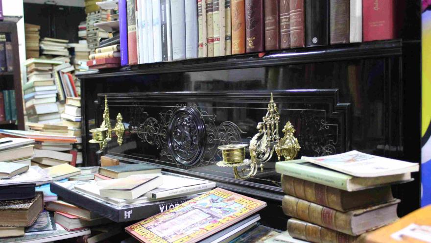 Piano antiguo con candelabros decorativos, rodeado de pilas de libros en una librería.